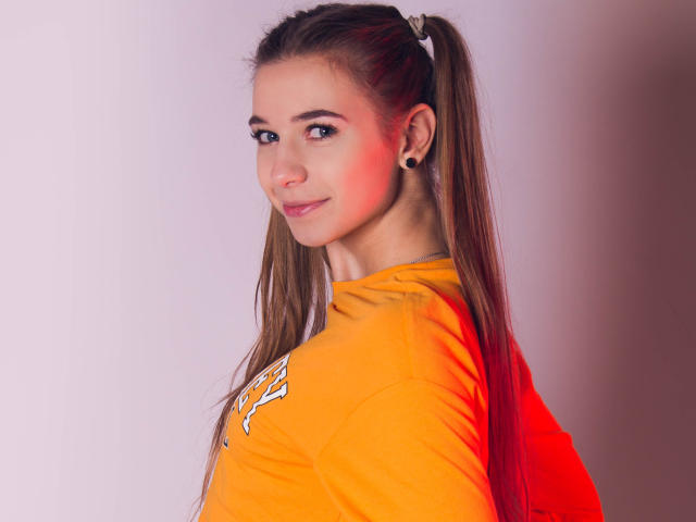 LucyBestCandy, with long brown pigtails, small black ear studs, and a cute smile, wearing an orange shirt, has a red highlight on her cheek.