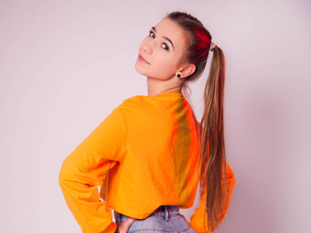 LucyBestCandy with long brown ponytail, wearing an orange long-sleeve shirt, blue jeans, and small earrings, looking over her shoulder with a slight smile.