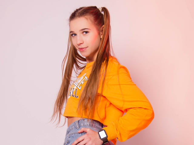 LucyBestCandy with long brown pigtails, orange crop top, and blue jeans, showing her ass with a hand on her hip, wearing a black smartwatch.