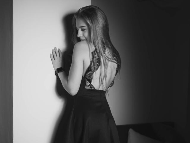 LucyBestCandy with long blonde hair, wearing a black lace-back dress, standing against a wall, hands up, looking down, black wristband, black and white photo, soft light.