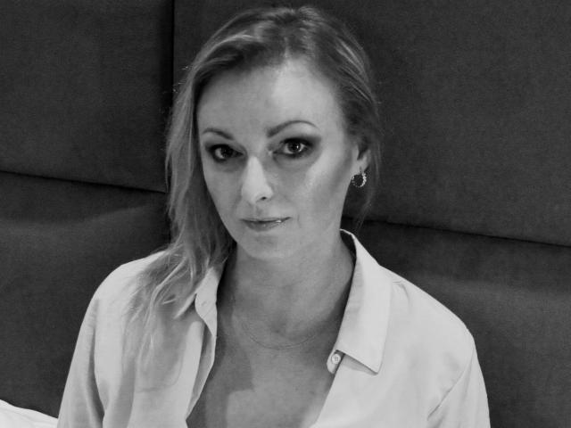 Black-and-white photo of XLovelyVanessa with blonde hair, wearing an open shirt, showing small tits, and a subtle smile, wearing small earrings, looking at the camera.