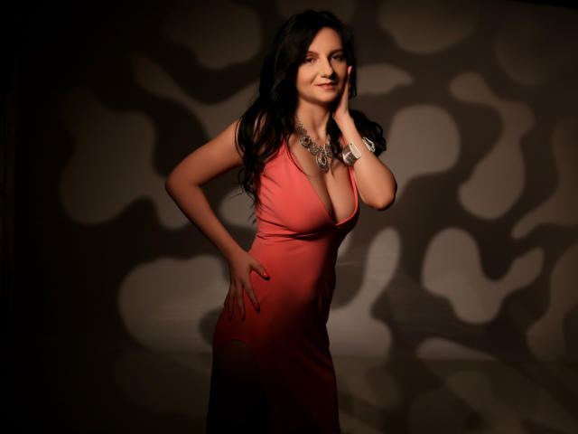 MadameAlexaX with long black hair, wearing a red dress with a deep cleavage, touching her face, hand on hip, necklace, shadow pattern on wall, smiling.
