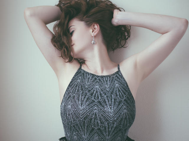 ChicEva with arms raised, wavy brown hair, wearing a sparkly black halter top, showcasing her tits and slim arms, wearing dangling earrings, in a soft light.