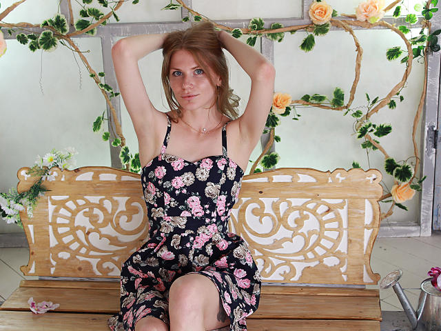 GretyArdent with arms up, wearing a black floral dress, sitting on a wooden bench with intricate carvings, looking at the camera with a slight smile.