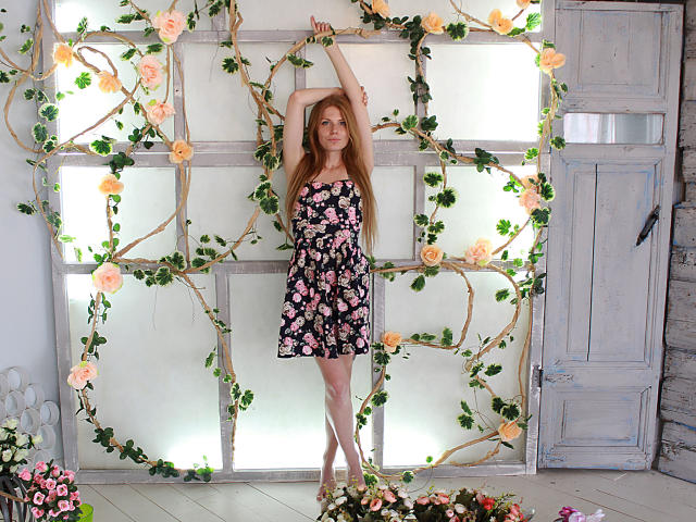 GretyArdent with long red hair, arms raised, wearing a black floral dress, standing in front of a vine-covered window, pale skin, and a cute, slender, sexy body.
