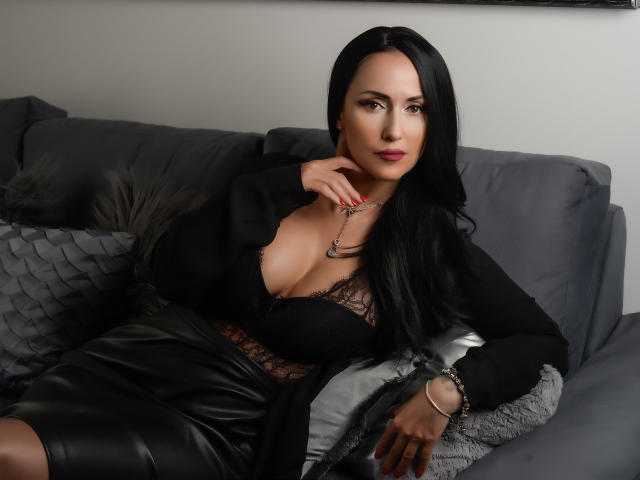 MissVonTease with black hair, wearing a black lace top and leather skirt, sitting on a gray couch, showcasing her big boobs and sexy cleavage, with a necklace.