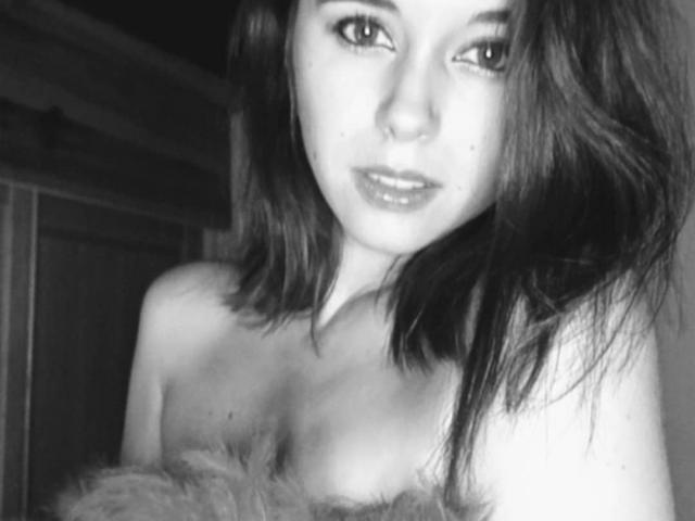 Black-and-white selfie of VirginieFrancaise with wavy dark hair, nose ring, and slightly parted glossy lips, showing her bare shoulders and a fluffy object in front of her.