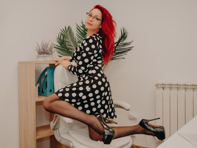 DirtyMature with red hair, glasses, black polka dot dress, black stockings, and black high heels, kneeling on white chair, looking over shoulder, ass raised.