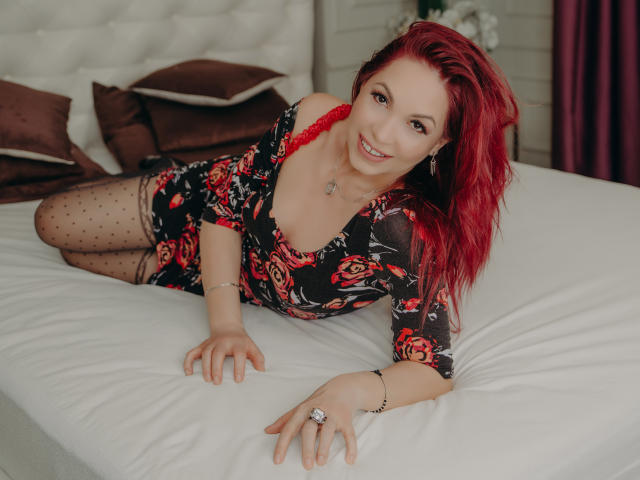 DirttyMature with red hair, wearing a floral dress, red lace bra, and polka dot tights, lying on a bed, smiling, showing her tits, and wearing a ring.