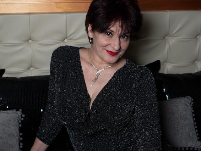 AlbertaAllen with short dark hair, red lipstick, and sparkly black dress, wearing a necklace, sitting on a black cushioned couch, smiling at the camera. Her tits are visible.
