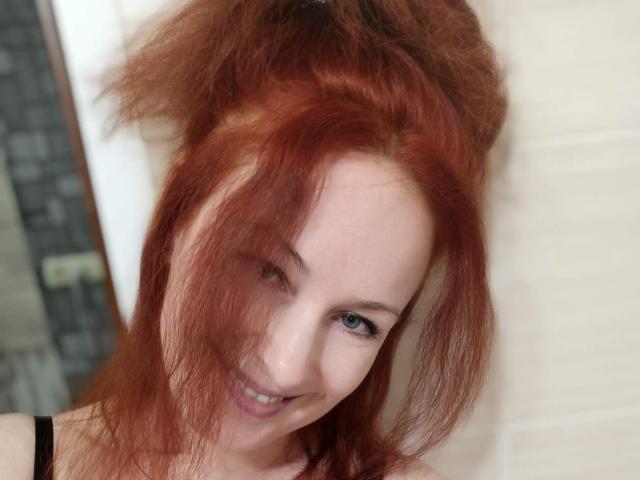 MarleneDavies with messy red hair, smiling, blue eyes, fair skin, black top, and a cute, slightly goofy look, leaning against a white door.