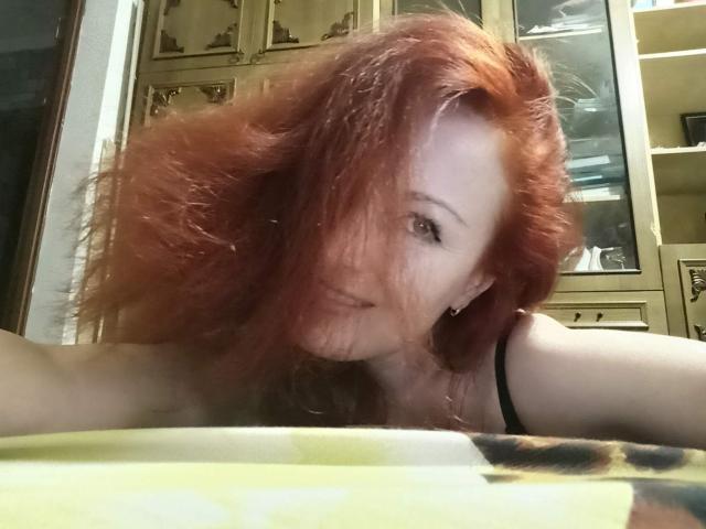 MarleneDavies with red, messy hair, black strap, and subtle smile, leaning on yellow and brown patterned surface, showing her pale skin and small nose.