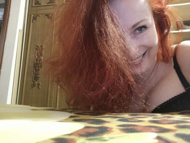 MarleneDavies with red hair, smiling at the camera, wearing black lace bra, showing her tits, leaning forward, on a leopard print blanket, wooden doors in background.