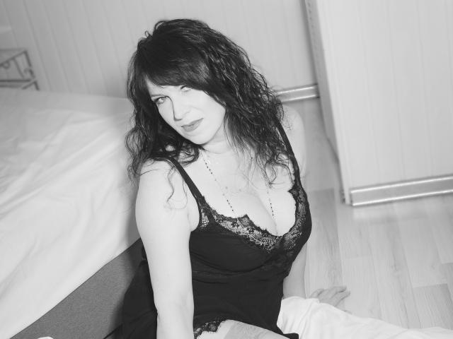 KellyMatureX with wavy black hair, wearing a black lace camisole, sitting on the floor, showing her tits, next to a bed with white sheets, in a black-and-white photo.