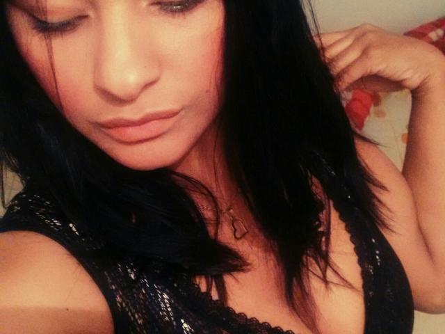 KarlysHoney with black lace bra, showing her tits, has dark hair, and a subtle pout, wearing a necklace, in a slightly blurred selfie.