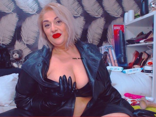 SexyyMilf, blonde with red lipstick, wearing black latex outfit, kneeling on bed, showing her ass, black boots, smiling, sex toys in background, patterned wallpaper.