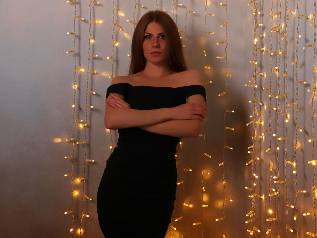 GretyArdent stands in a black off-shoulder dress, arms crossed, with long brown hair, glowing fairy lights in the background, and a subtle, confident smile.