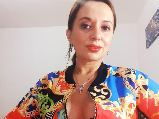 MaitresseBelle heeft bruin haar, roze oogschaduw, rode lippenstift en draagt een kleurrijke, versierde jas met een diepe V-hals, waardoor haar borsten zichtbaar zijn. Ze draagt een zilveren ketting..