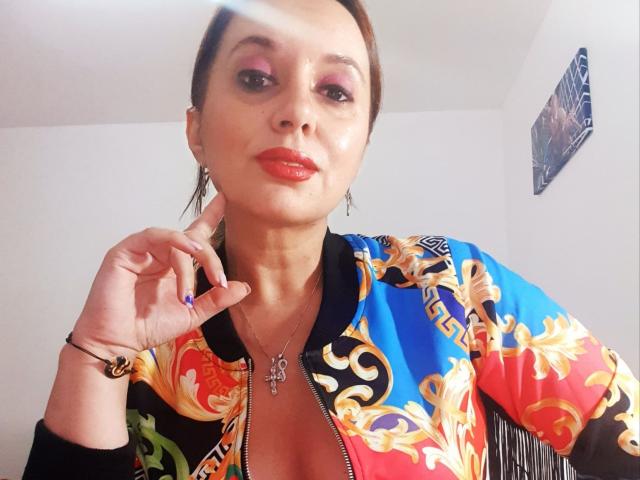 MaitresseBelle with red lipstick, pink eyeshadow, and brown hair in a ponytail, wearing a colorful, floral-patterned jacket, zipped down to show cleavage, holding her chin.