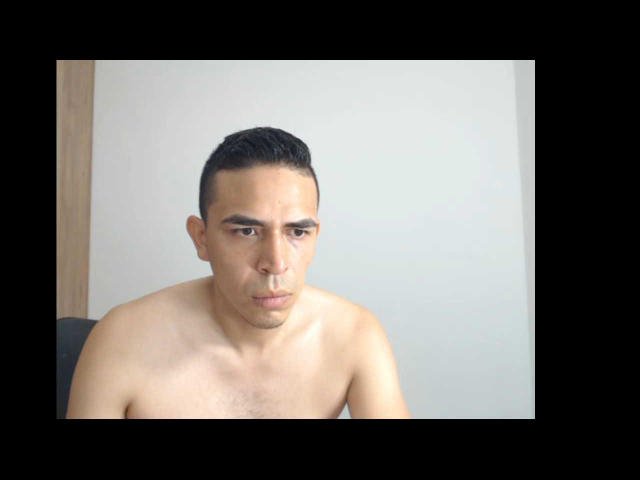 JeanCarlos is shirtless, with short black hair and a serious expression, looking to the side. His small, dark nipples are visible on his light brown chest.