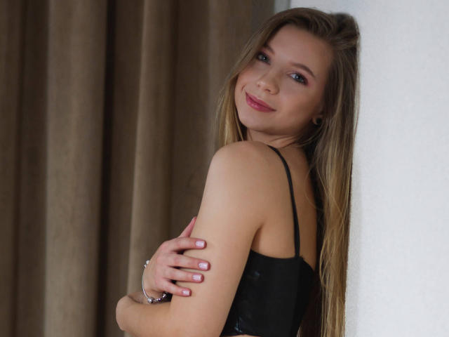 LucyBestCandy with long blonde hair, black lace top, arms crossed, smiling at camera, pink nails, soft light, white wall, brown curtain, relaxed, sexy, subtle makeup.