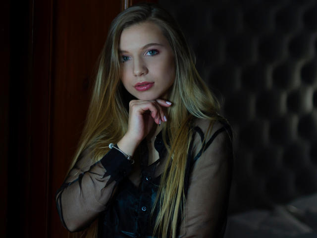 LucyBestCandy with blonde hair, blue eyes, and pink lips, wearing a sheer black top, rests her chin on her hand, showing off her tits and a sexy smile.