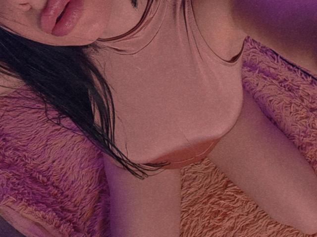 MaxSandra's pink tank top, showing her tits, with black hair, pink lips, and purple-lit skin, sitting on a fluffy pink blanket. Her pussy is slightly visible.