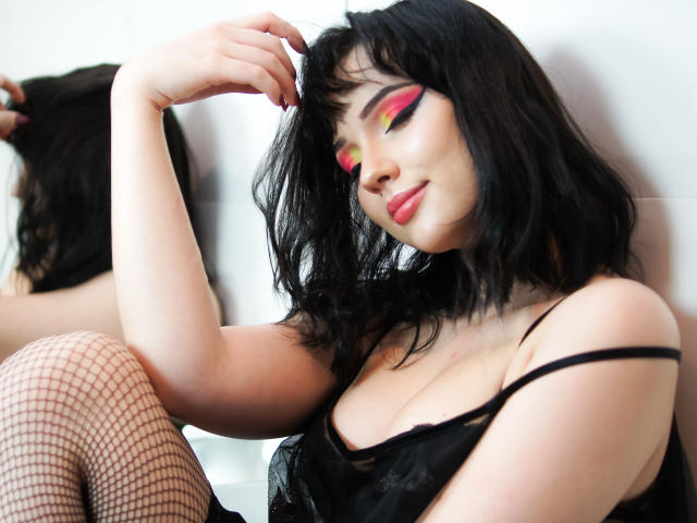 DeniseKitty with black hair, pink and yellow eye makeup, black lingerie, and fishnet stockings, sitting with a hand in her hair, showing her tits and a slight smile.