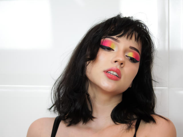 DeniseKitty with black hair, pink and yellow eye makeup, and glossy pink lips, slightly drooling, wearing a black top, has a sexy, seductive expression, and pale skin.