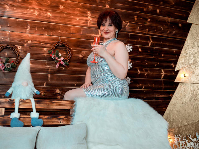 AlbertaAllen in a sparkly blue dress, sitting on a white fur-covered couch, holding a champagne glass, smiling, with a small gnomes and wooden wall in the background.