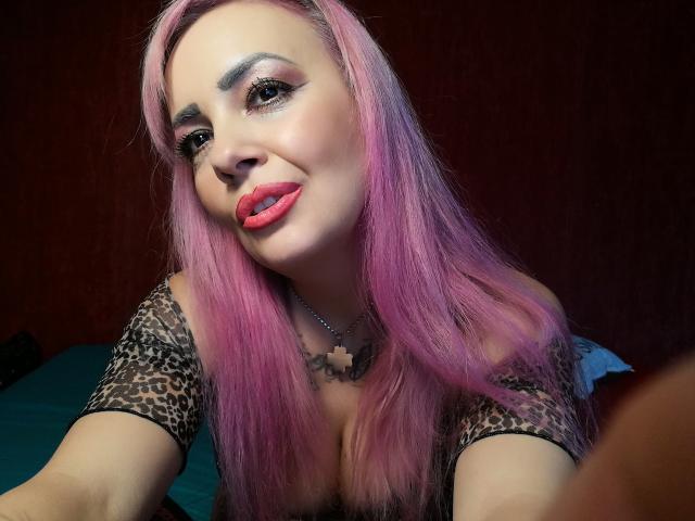 HotAdelayde69 with pink hair, leopard print top, red lips, and a cross necklace, smiling at the camera, showing her tits. She has dark eye makeup and a cute tattoo.