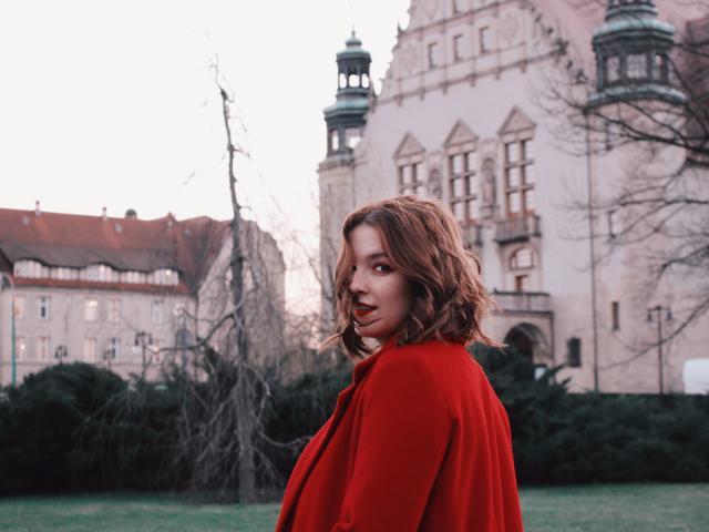 KenzieONEx with wavy brown hair, red coat, green grass, and a historic building in the background, looking over her shoulder with a slight smile.