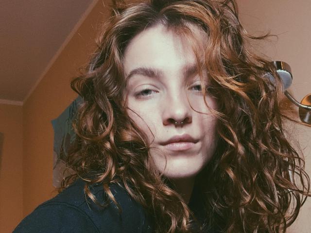 KenzieONEx with wavy, dirty-brown hair, pale skin, nose ring, and green eyes, wearing a dark top, looking slightly to the side with a neutral expression.