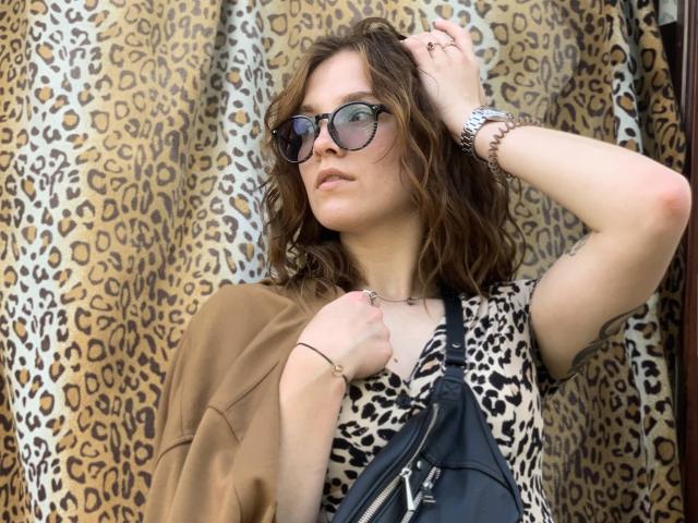 KenzieONEx has wavy brown hair, wears dark sunglasses, a leopard print top, and a brown jacket, with a black bag, a silver watch, and a bracelet.