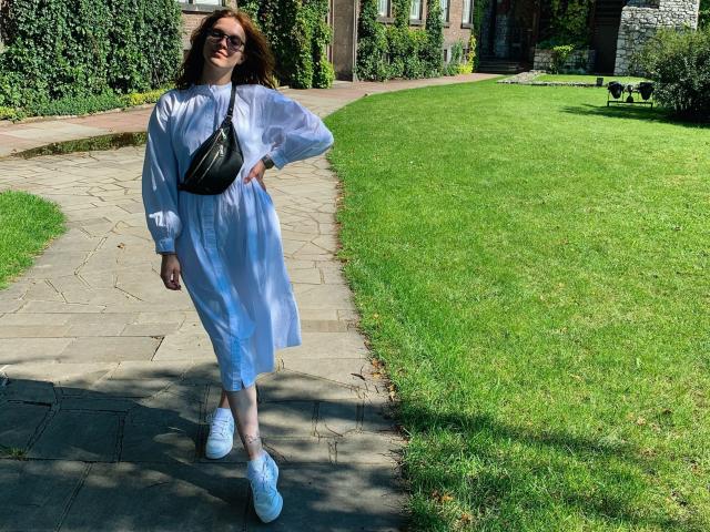 KenzieONEx with red hair, glasses, blue dress, black crossbody bag, white sneakers, standing on stone path, green lawn, and brick building in background, smiling.