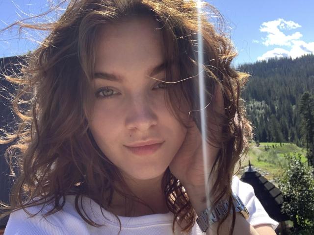 KenzieONEx with curly brown hair, green eyes, and a slight smile, wearing a white shirt, has a sunbeam in her face, and a hand touching her cheek.