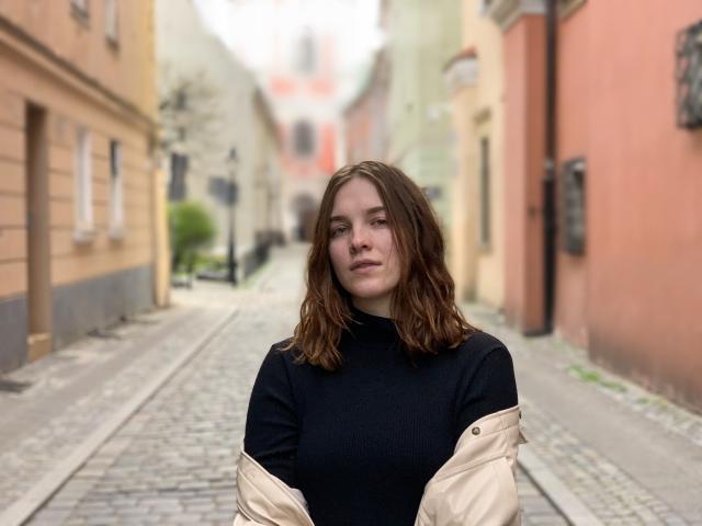 KenzieONEx with wavy brown hair, wearing a black turtleneck and beige jacket, standing on a cobblestone street, looking slightly off-camera with a neutral expression.