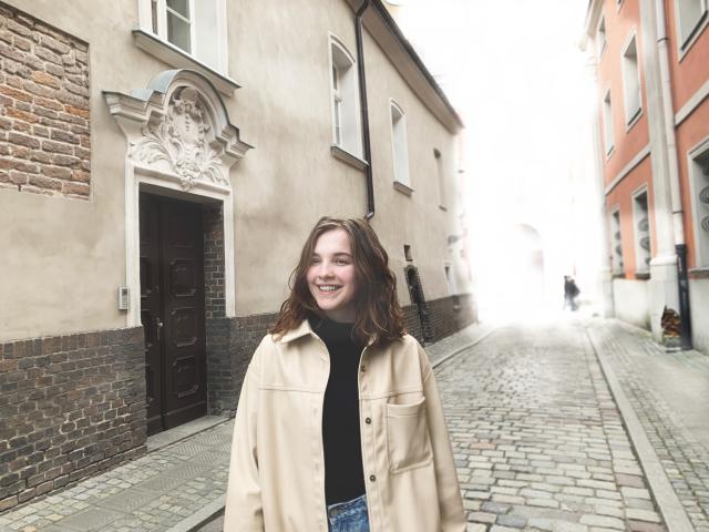 KenzieONEx has wavy brown hair, a smiling face, wears a beige coat over a black turtleneck, and blue jeans, standing on a cobblestone street.