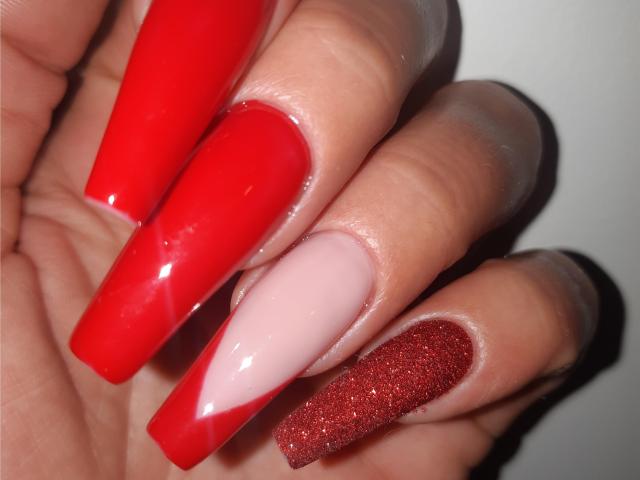 ExquisDunya's hand with long, shiny red nails on two fingers, pale pink nail on middle finger, and red glitter nail on pinky finger, showcasing a sexy, colorful manicure.