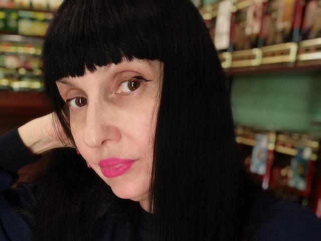 MelanyBanks with black straight hair, brown eyes, pink lipstick, and a hand in her hair, wearing a navy shirt, in a blurry store background. Her nose is slightly upturned.
