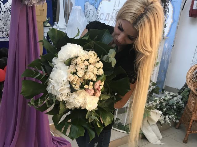 Blond AlisaAleksandr with long hair, holding a bouquet of white roses and green leaves, smiling, wearing a black top, in a floral shop. Boobs not visible.