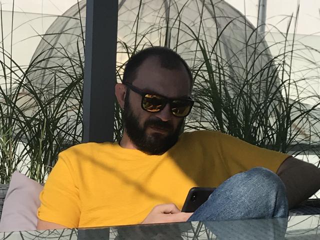 Bearded dude with dark sunglasses, yellow shirt, and blue jeans, holding phone, leaning back with a relaxed, slightly dirty look, surrounded by green plant shadows.