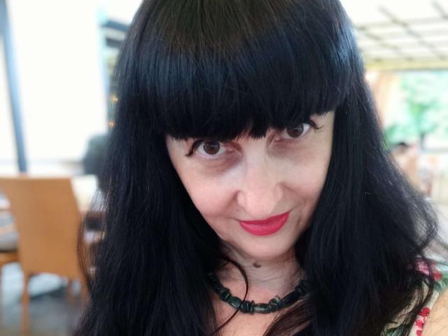 MelanyBanks has black, straight, shoulder-length hair with bangs, brown eyes, red lipstick, and a black beaded necklace. Her face is slightly smiling with a small mole on her chin.