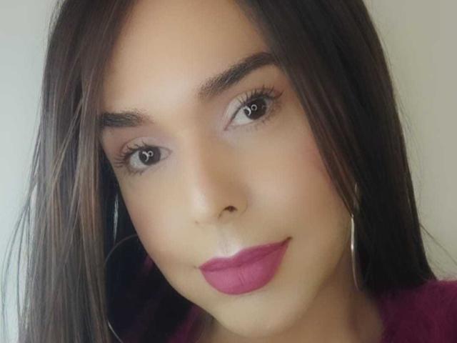Le visage de Jhovanna, avec ses cheveux bruns foncés, son rouge à lèvres rose, ses longs cils et ses petits boucles d'oreilles, affiche un sourire subtil. Sa peau est bronzée et elle a des sourcils bien dessinés..