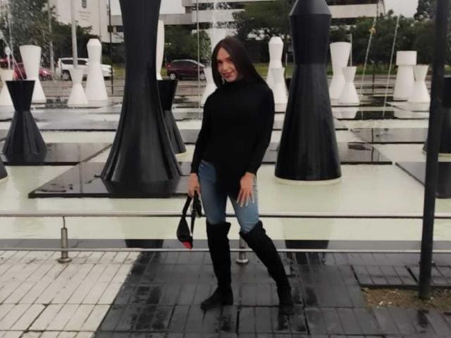 Jhovanna, con un seno di media grandezza, indossa un maglione nero, jeans blu e stivali neri che arrivano al ginocchio, tiene in mano una borsa nera e si trova di fronte a sculture alte in bianco e nero..