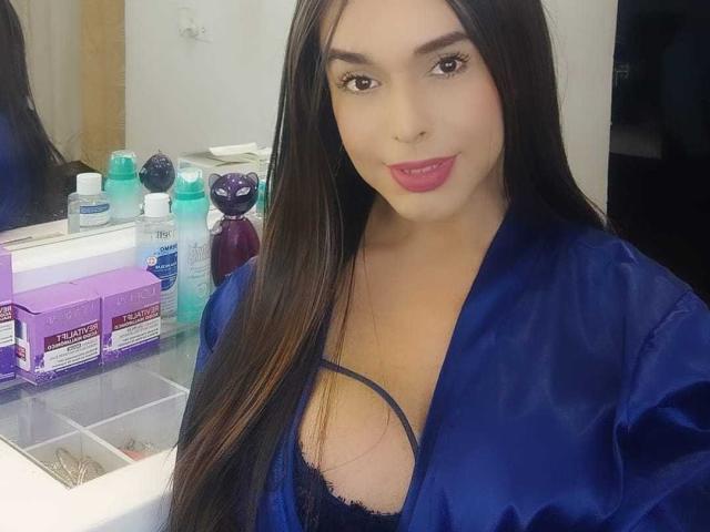 Jhovanna, con i suoi lunghi capelli neri, indossa una veste di raso blu che mette in mostra il suo seno generoso, coperto da un reggiseno di pizzo nero, sorride con il rossetto rosa e si trova di fronte a un bancone per il trucco..