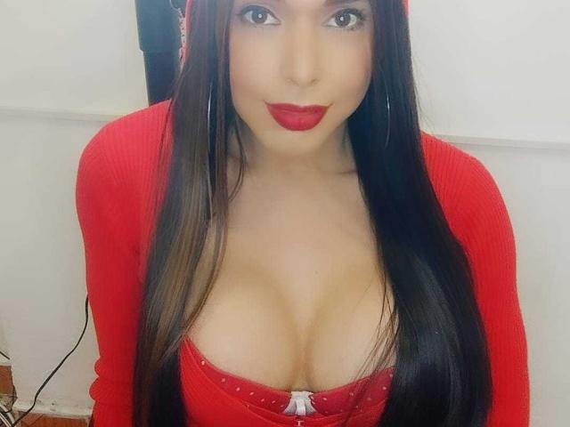 Jhovanna, con i suoi lunghi capelli neri, un cardigan rosso e il rossetto rosso, mostra il suo seno prosperoso in una maglia rossa con un fiocco bianco. Sta guardando la fotocamera con un sorriso..