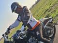 FrancaiseAnnaDom on a black motorcycle, wearing a helmet, reflective visor, black and white jacket, orange vest, black pants, and gloves, leaning forward on a road.
