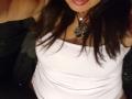 HotSensationX with dark hair, wearing a white tank top, showing her tits, and a leaf pendant necklace, with her tongue slightly out, in a dimly lit setting.