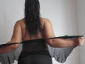SaimaJaye69 with black wavy hair, back to camera, removing sheer black polka-dot top, revealing bare back and black pants, holding top with both hands.